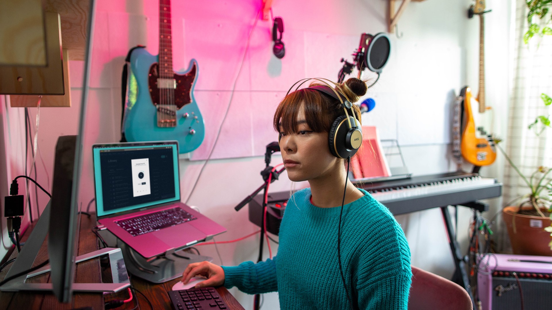 An independent artist in a teal sweater and studio headphones works at a home setup, editing a track on a laptop showing a music library, with electric guitars, a bass, and a keyboard on the wall behind them.